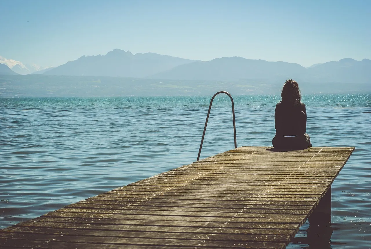 A person sitting at the end of a wooden dock, looking out across the water.
