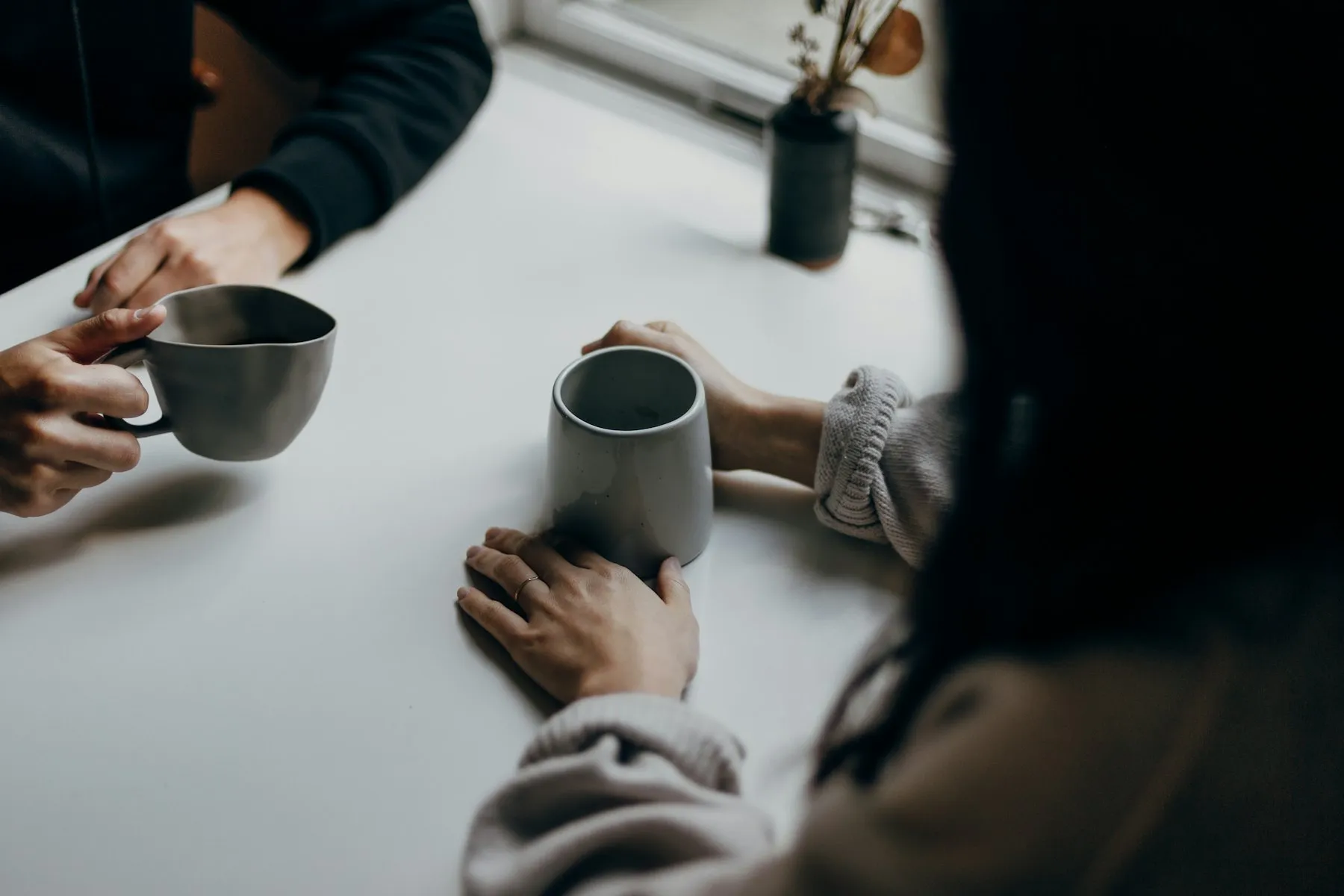 Two people holding mugs across a table, mid-conversation.