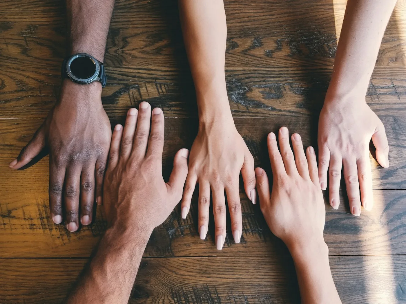 Five hands resting side by side on a wooden table.
